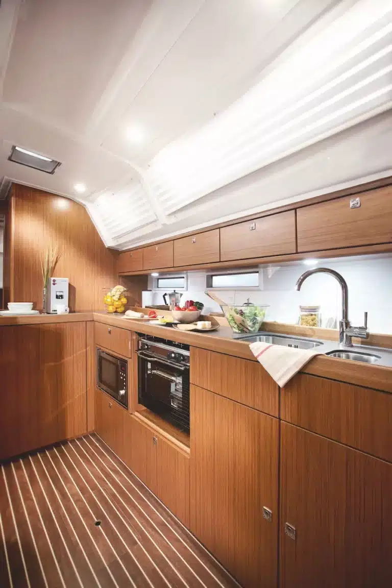 Eine moderne Yachtküche mit Holzschränken, Geräten aus Edelstahl und einer eleganten Arbeitsplatte. Sonnenlicht fällt durch das Oberlicht ein und erhellt den Raum der Segelyacht.