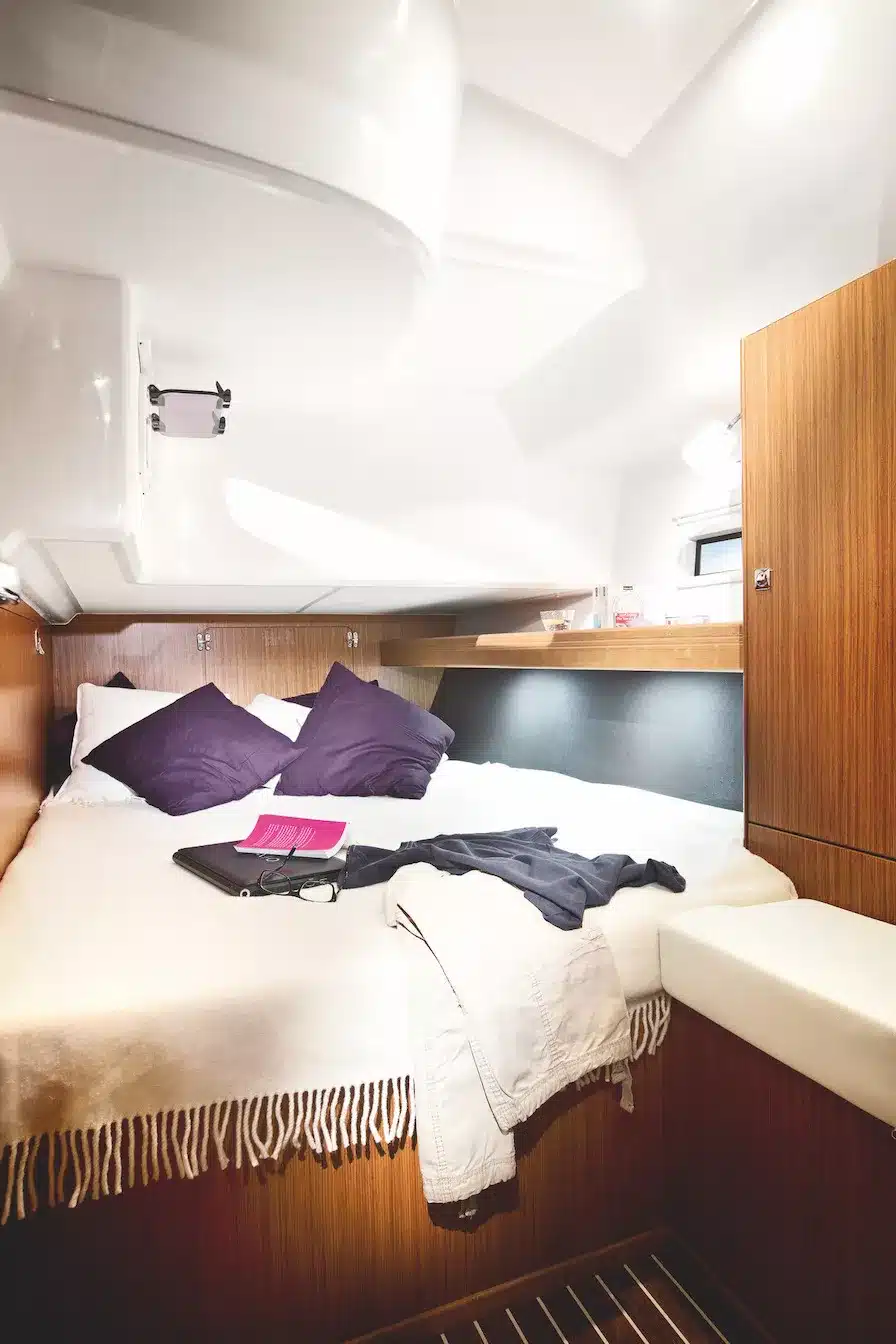 Innenansicht einer gemütlichen Segelyachtkabine mit einem ordentlich gemachten Bett mit weißer Bettwäsche und violetten Kissen, einem Holzschrank und Kleidungsstücken auf dem Bett.