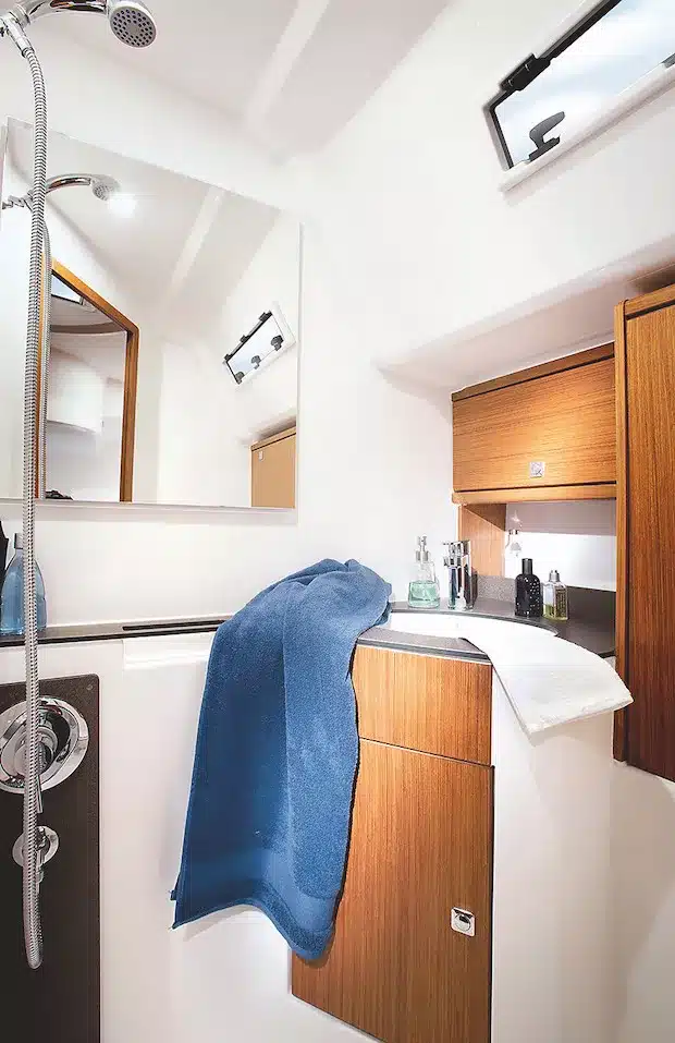 Modernes, kompaktes Bootsbadezimmer mit Holzschränken, einem weißen Waschbecken, einer Dusche, einem Spiegel und einem links hängenden Handtuch, das die effiziente Nutzung des begrenzten Raums während eines Segeltörns darstellt.