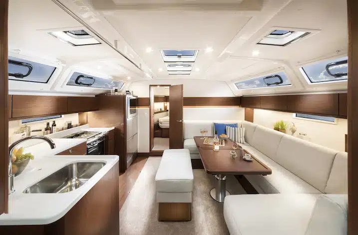 Innenansicht einer Luxusyacht mit elegantem Küchenbereich, gemütlichem Essbereich mit cremefarbenem Sofa und hölzernem Couchtisch, beleuchtet durch natürliches Licht aus mehreren Luken an der Decke während eines Segelabenteuers