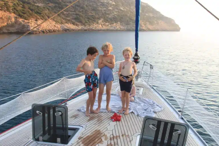 Drei Kinder stehen lächelnd auf dem Deck einer Segelyacht, umgeben von ruhigem blauen Wasser und einer felsigen Küste bei Sonnenuntergang.