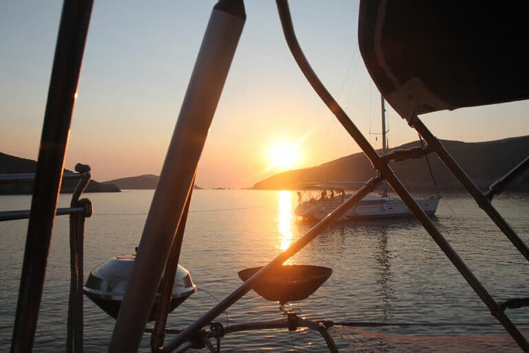 Ein ruhiger Sonnenuntergang von einem Katamaran aus. Segelboote sind auf ruhigem Wasser zwischen sanften Hügeln zu sehen, eingerahmt von Bootstakelage und einem Rettungsring, und die Sonne wirft einen goldenen Schein.