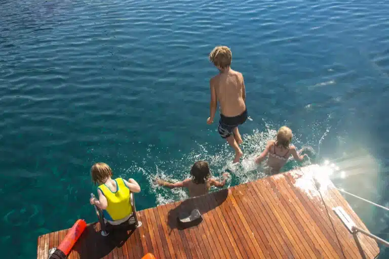 Eine Gruppe Kinder springt an einem sonnigen Tag freudig vom Holzdeck einer Segelyacht ins Meer.