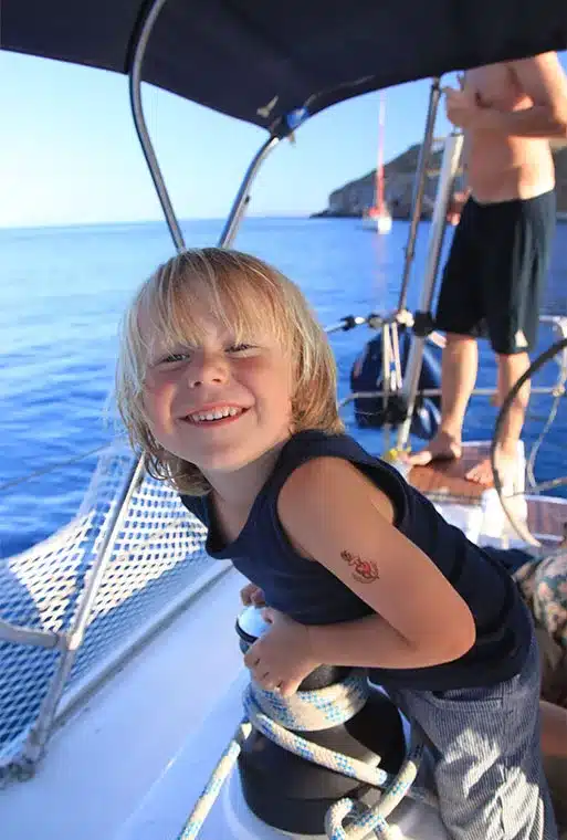 Ein fröhlicher kleiner Junge mit blondem Haar lächelt breit auf einem Segelboot während eines Segelurlaubs, mit klarem blauen Himmel und Meer im Hintergrund, während eine andere Person teilweise sichtbar hinter ihm steht.