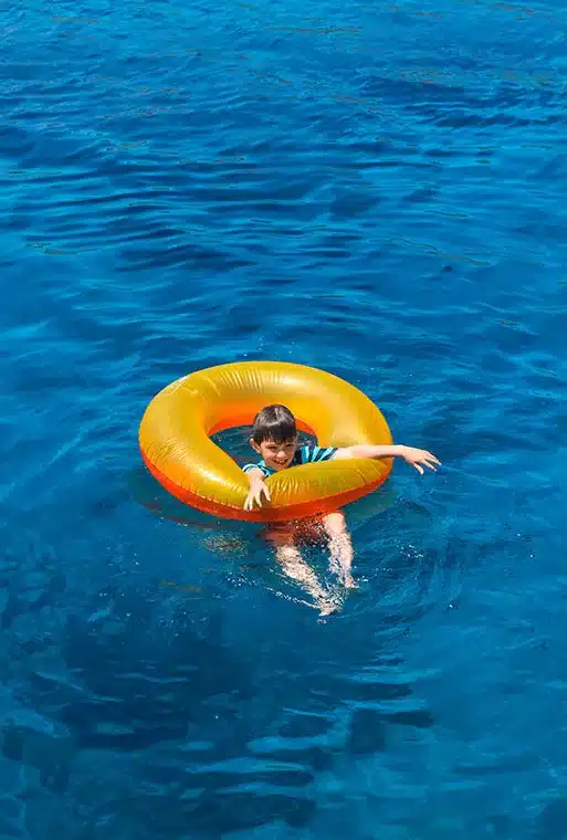 Ein Kind in einem gestreiften Hemd schwimmt auf einem leuchtend orangefarbenen aufblasbaren Ring in einem klaren blauen Ozean in der Nähe einer Segelyacht.