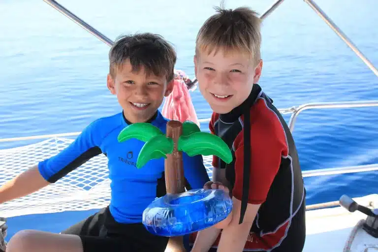 Zwei Jungen mit freudigem Gesichtsausdruck und Schwimmwesten halten eine aufblasbare Palme auf einem Boot, während sie unter klarem blauen Himmel segeln.