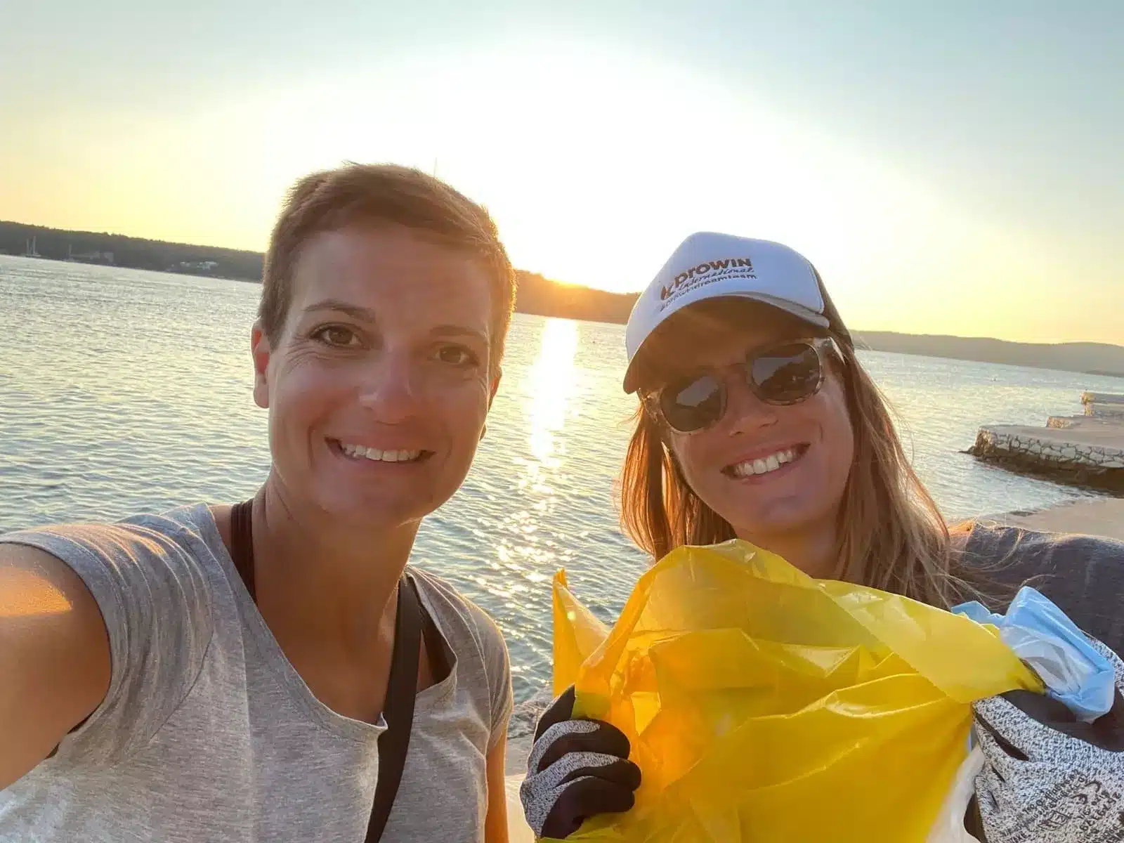 Zwei Frauen lächeln für ein Selfie mit einem See im Hintergrund bei Sonnenuntergang. Eine trägt eine Mütze und eine Sonnenbrille und hält einen gelben Müllsack in der Hand, möglicherweise nach einer Reinigungsaktion beim Segeln.