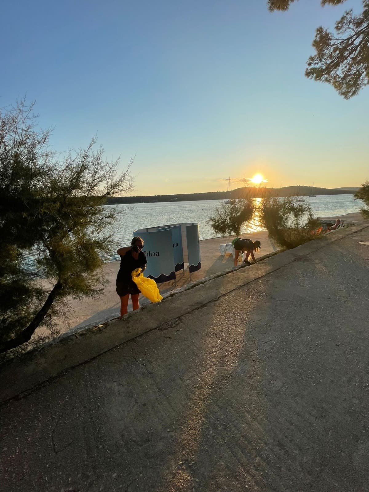 Eine Person geht bei Sonnenuntergang einen Weg am Seeufer entlang und trägt eine gelbe Tasche, während sich eine andere Person in der Nähe bückt. Der See reflektiert das Sonnenlicht, eingerahmt von Bäumen und einem klaren Himmel, was an