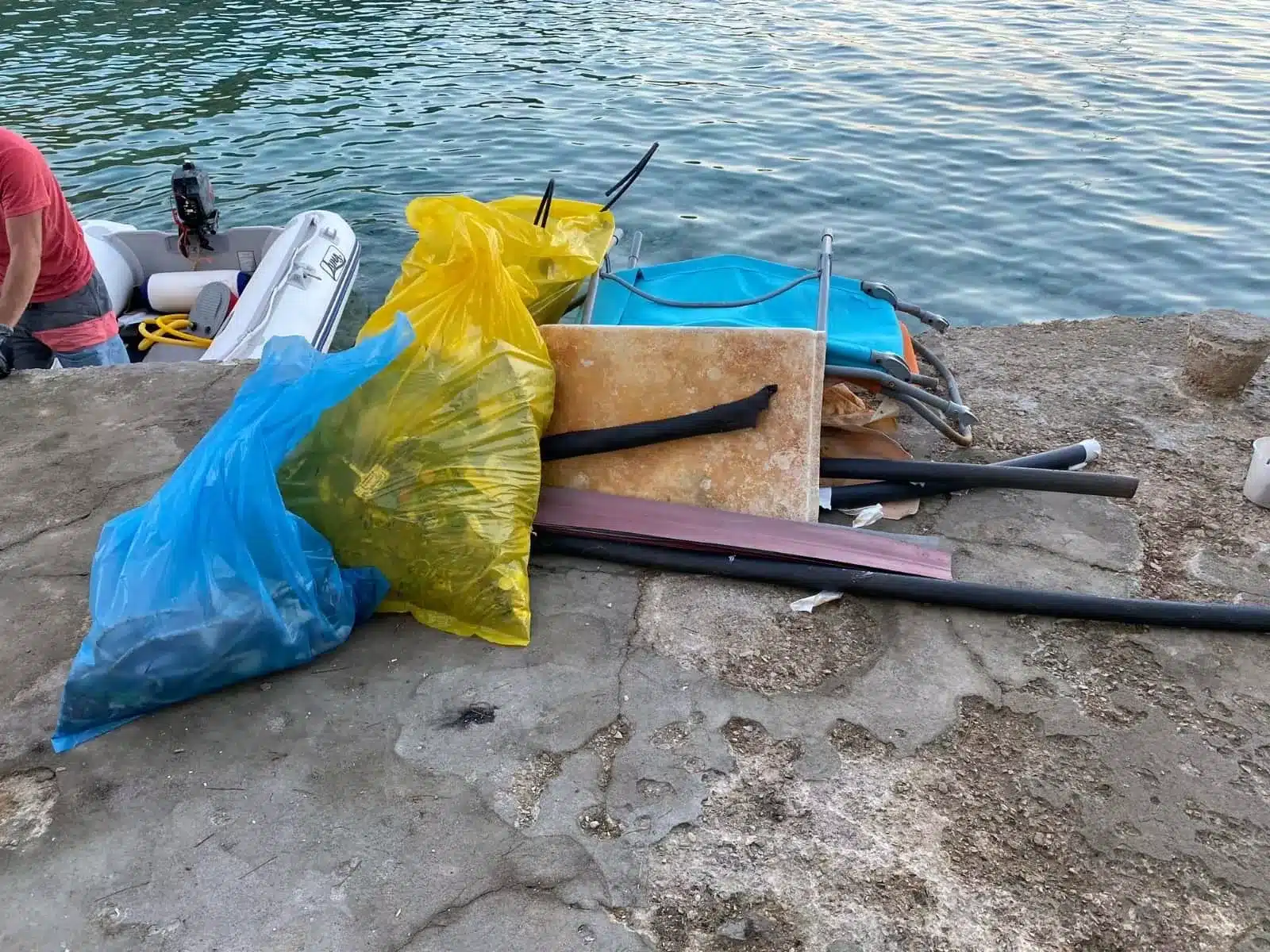Ein Haufen verschiedener Abfälle, darunter Holzbretter, blaue und gelbe Plastiktüten und Metallstangen, auf einem Betonsteg neben einem Gewässer mit einer kleinen Segelyacht in der Nähe.