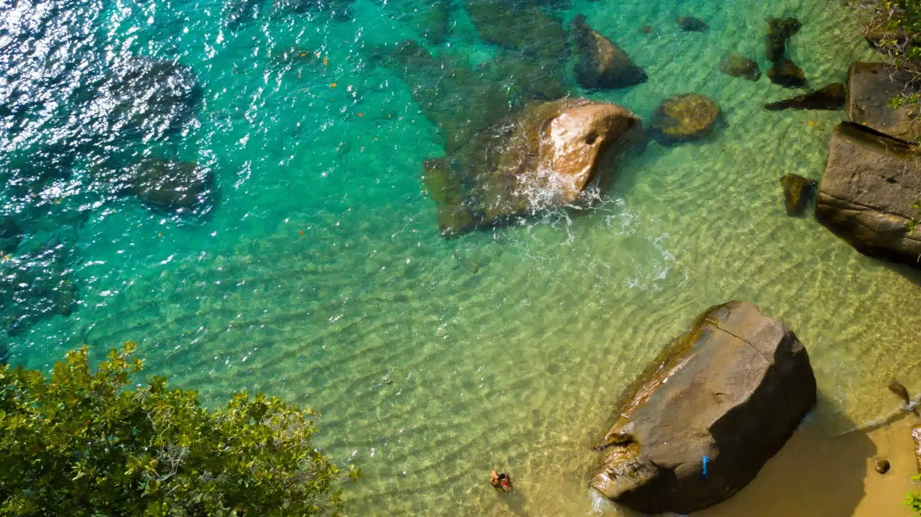 Luftaufnahme eines klaren türkisfarbenen Meeres mit großen Felsen in Ufernähe und ein paar Menschen, die während ihres Segelurlaubs um sie herum schwimmen und schnorcheln. Grünes Laub umrahmt teilweise den oberen Rand des