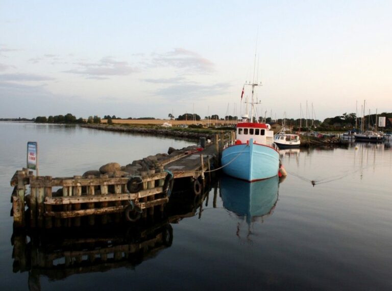 Eine ruhige Hafenszene mit einer blau-roten Yacht, die an einem Holzsteg festgemacht ist und sich in der Abenddämmerung im ruhigen Wasser spiegelt. Im Hintergrund sind Grasfelder zu sehen.