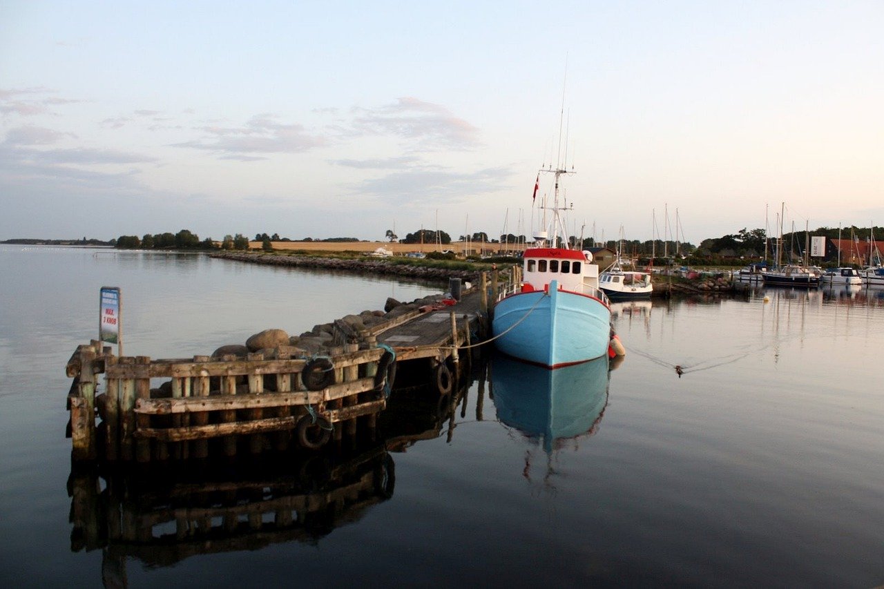 Eine ruhige Hafenszene in der Abenddämmerung mit einem blauen Boot, das an einem Holzsteg festgemacht ist, und in dessen ruhigem Wasser sich der Himmel und die umgebende Landschaft spiegeln, bereit für einen Segeltörn.