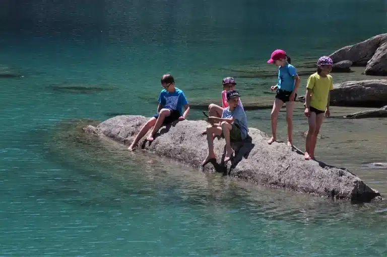 Vier Kinder auf einem Felsen an einem See tauchen ihre Füße ins Wasser, während zwei weitere Kinder hinter ihnen stehen, alle in Sommerkleidung zum Segeln gekleidet.