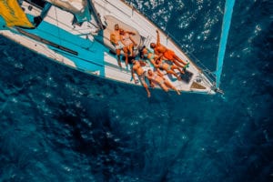 Luftaufnahme einer Gruppe von Menschen, die auf dem Deck einer Segelyacht mit blauem Segel ein Sonnenbad nehmen und auf dem leuchtend blauen Ozeanwasser treiben.