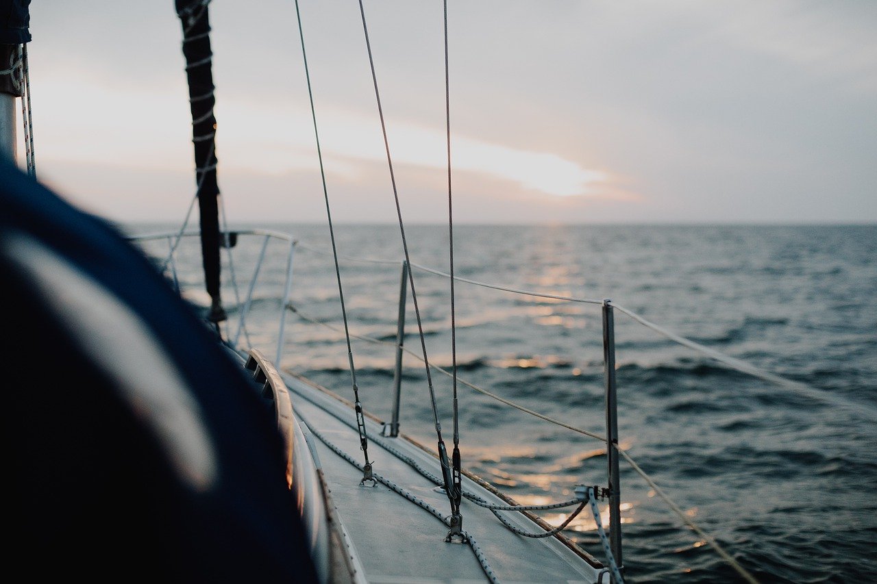 Blick vom Deck einer Segelyacht auf das Meer während des Sonnenuntergangs, mit Fokus auf Reling und Takelage, mit ruhigem Ozeanwasser und sanftem Himmel im Hintergrund.