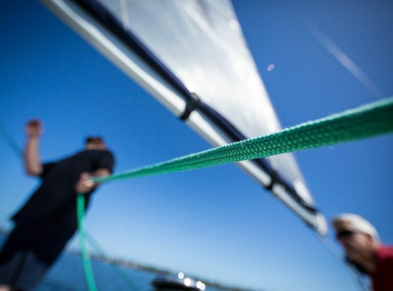 Eine Nahaufnahme eines grünen Seils auf einer Segelyacht mit unscharfen Bildern von zwei Personen, die unter einem klaren blauen Himmel segeln.