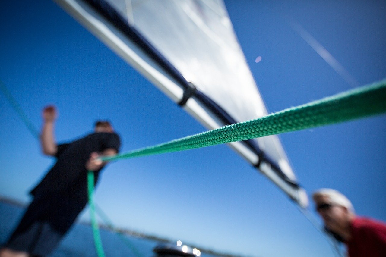 Unscharfes Bild von zwei Personen auf einer Segelyacht mit Fokus auf einem grünen Seil im Vordergrund; klarer blauer Himmel und Wasser im Hintergrund.