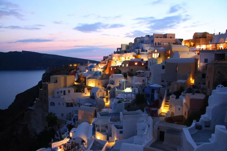 Die Dämmerung senkt sich über Oia, Santorini, mit seinen ikonischen weiß getünchten Häusern und blauen Kuppeln, die vor einem rosa und blauen Himmel erleuchtet sind, mit Blick auf das ruhige Meer, perfekt für einen ruhigen