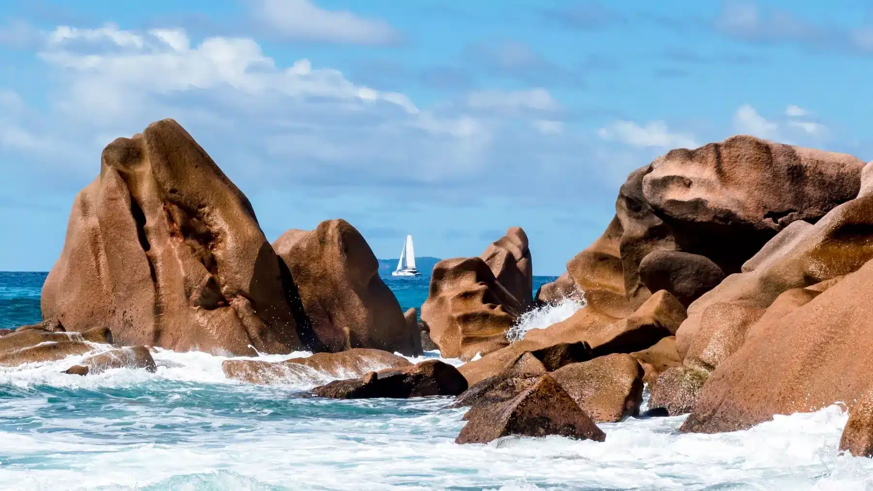 Wellen schlagen gegen große, glatte Felsbrocken an einer sonnenbeschienenen Küste. Unter klarem Himmel ist am blauen Ozeanhorizont eine einsame Segelyacht zu erkennen.