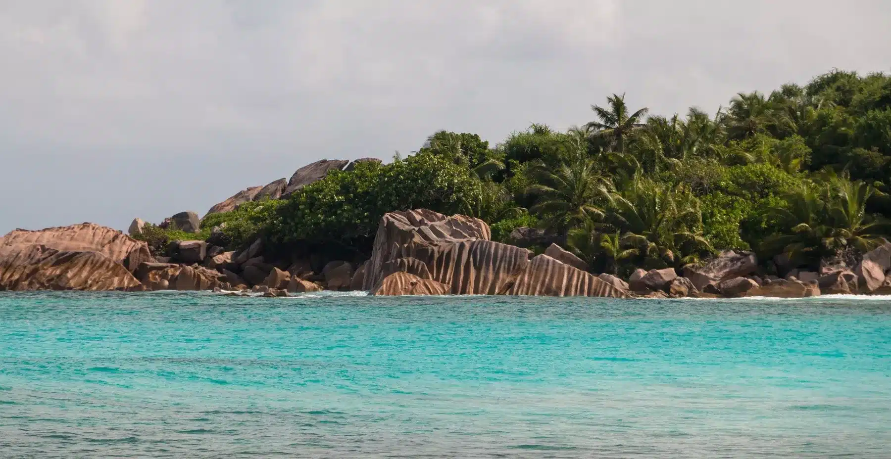 Eine ruhige Strandszene mit aquamarinblauem Wasser im Vordergrund und einer üppigen, grünen Insel mit markanten Felsvorsprüngen im Hintergrund unter einem klaren Himmel, perfekt für einen Segeltörn.