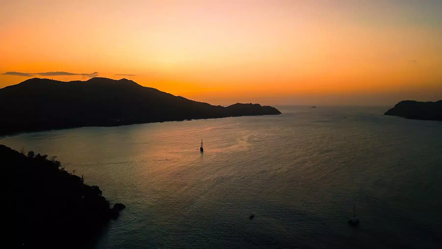 Luftaufnahme eines ruhigen Meeres bei Sonnenuntergang mit der Silhouette von Hügeln am Horizont und verstreuten Segelbooten auf dem Wasser, die die dunkelorangen Farbtöne des Himmels reflektieren.