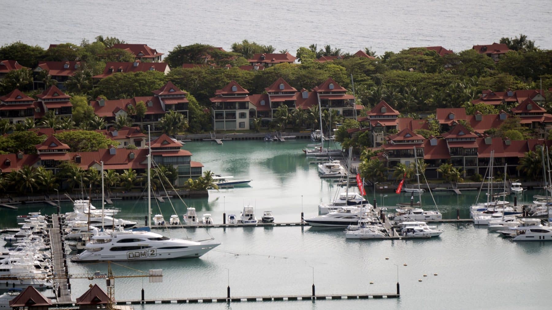 Luftaufnahme eines tropischen Yachthafens mit ordentlich geparkten Yachten, umgeben von Gebäuden mit roten Dächern und üppigem Grün, gelegen an einer ruhigen, blauen Bucht, perfekt für einen Segelurlaub