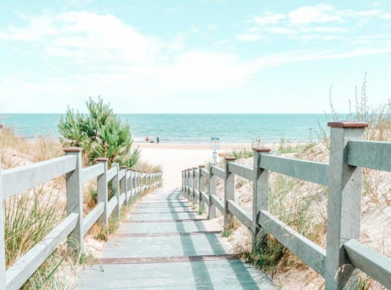 Ein malerischer Holzsteg mit weißem Geländer führt zu einem ruhigen Strand mit klarem blauen Himmel, einigen Menschen in der Ferne und einem ruhigen Meer. Gras und kleine Sträucher säumen den Weg zu