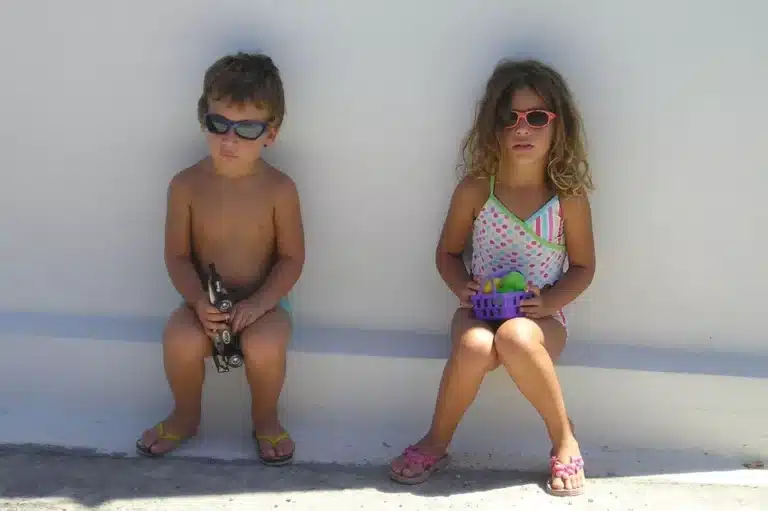 Zwei kleine Kinder sitzen vor einer weißen Wand. Der Junge links trägt eine Sonnenbrille und hält eine Spielzeug-Segelyacht, und das Mädchen rechts trägt eine Sonnenbrille, einen Badeanzug und hält eine