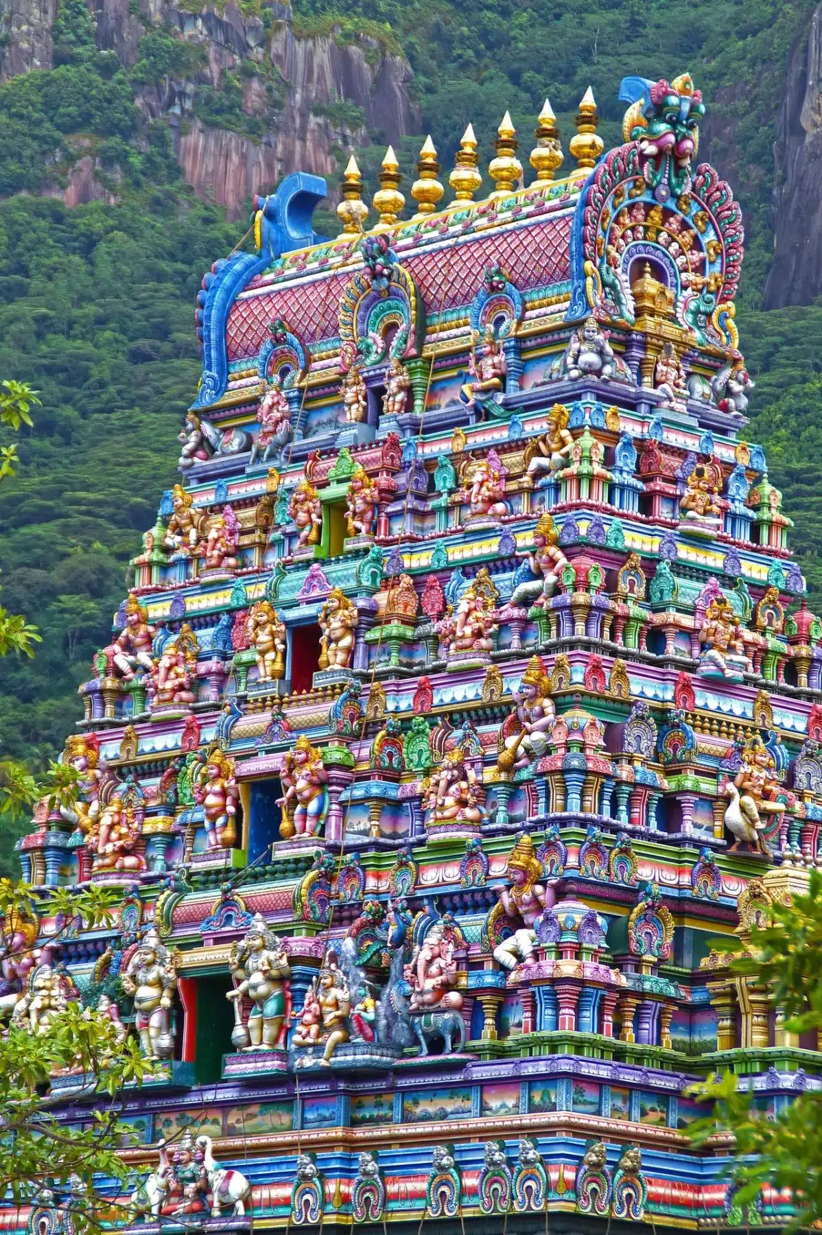Lebendiger und kunstvoll verzierter Turm eines Hindutempels, geschmückt mit farbenfrohen Statuen und Skulpturen von Gottheiten, vor der Kulisse üppiger grüner Berge, der an die Ruhe eines Seg erinnert