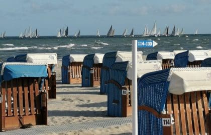 Strandszene mit Reihen überdachter Strandkörbe im Vordergrund und zahlreichen Segelyachten auf dem Meer im Hintergrund unter klarem Himmel.