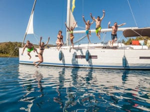 Eine Gruppe fröhlicher Menschen springt an einem sonnigen Tag während eines Segeltörns von einem Segelboot in einen See, im Hintergrund ist der Himmel klar.