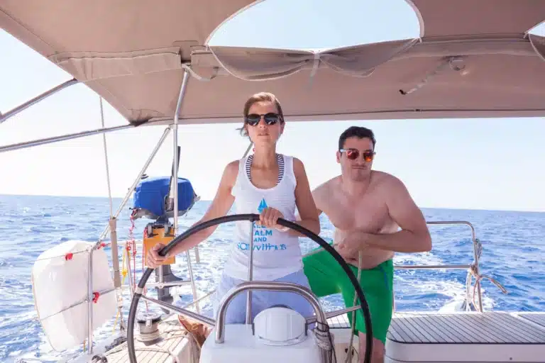 Zwei Personen stehen an einem sonnigen Tag unter einem Baldachin auf einem Segelboot und nehmen an einem Skippertraining teil. Die Frau am Steuer trägt eine Sonnenbrille und ein weißes Tanktop, der Mann ist mit freiem Oberkörper und grünen Shorts unterwegs. Im Hintergrund sind das Meer und ein klarer Himmel zu sehen.
