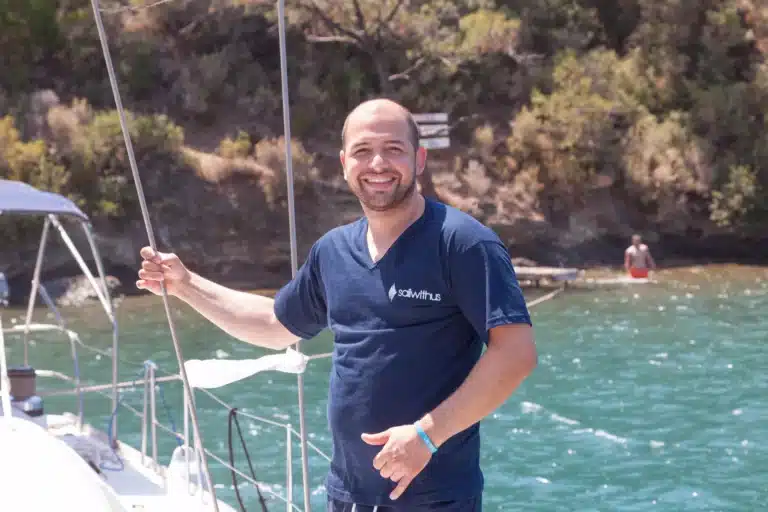 Ein lächelnder Mann in einem blauen T-Shirt steht auf einem Katamaran und hält sich mit einer Hand an einem Seil fest. Im Hintergrund sind ein ruhiges Meer und eine üppig grüne Küste zu sehen.