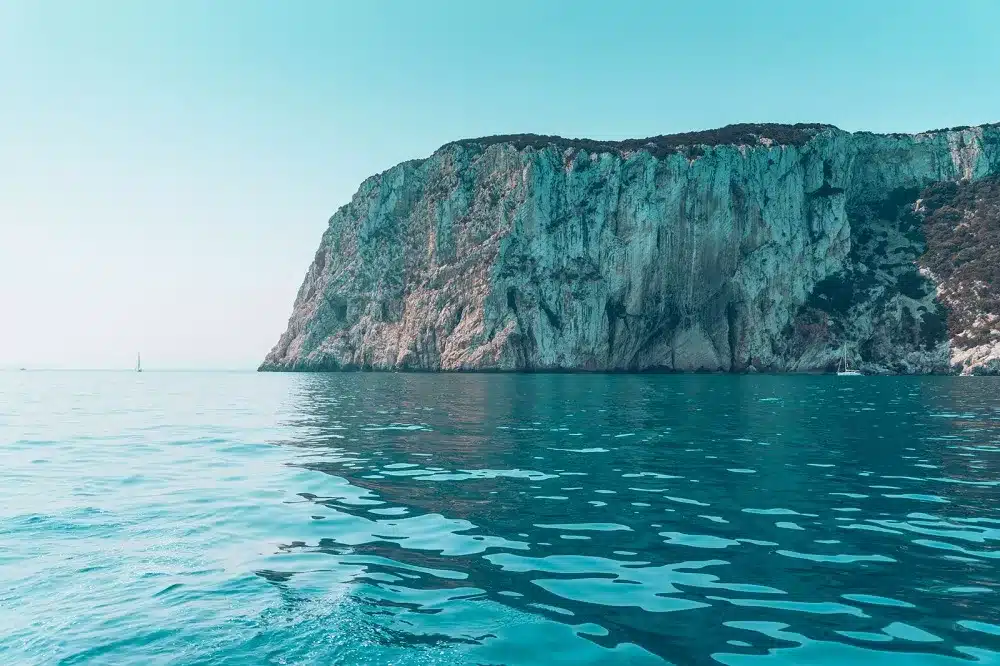 Eine ruhige Meereslandschaft mit einer großen, schroffen Klippe, die sich unter einem klaren Himmel aus blaugrünem Wasser erhebt. Die Oberfläche der Klippe ist mit grünem Laub und Felsflecken strukturiert und blickt auf eine Segelyacht, die sanft vorbeigleitet.