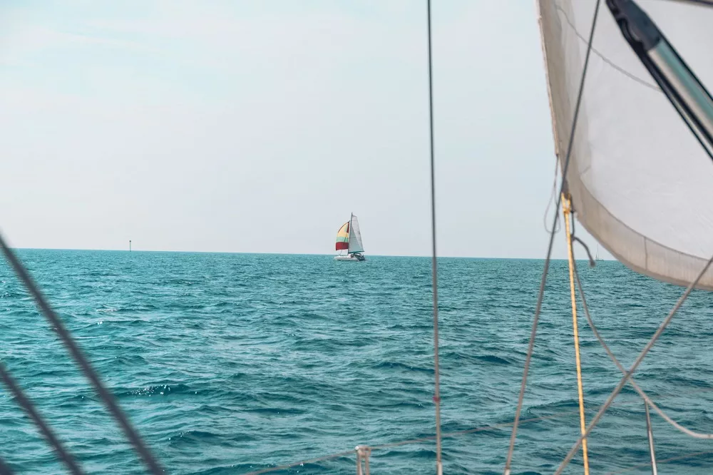 Blick von einem Katamaran. Auf der rechten Seite ist ein Teil des Segels zu sehen. In der Ferne ist ein weiteres Segelboot zu sehen, das auf einem ruhigen, blauen Meer unter einem klaren Himmel segelt.