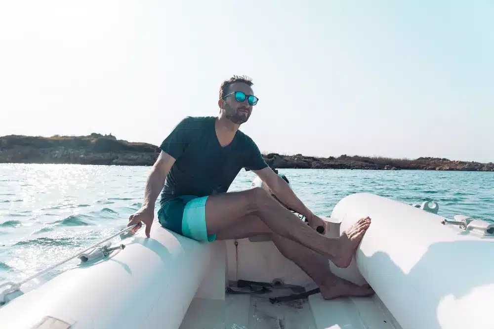 Ein Mann mit Sonnenbrille und Sommerkleidung sitzt entspannt auf der Kante einer Segelyacht, im Hintergrund der klare blaue Himmel und das ruhige Meer.