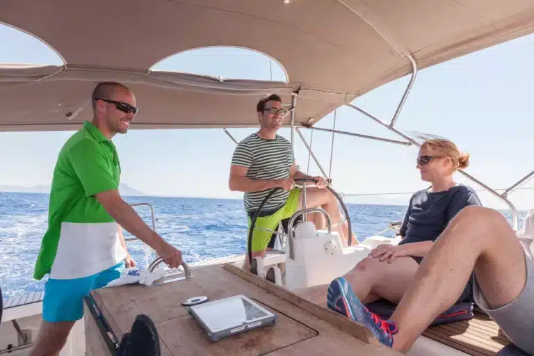 Drei Personen auf einer Segelyacht: eine Frau sitzt und zwei Männer, einer steuert. Sie unterhalten sich fröhlich unter einem Sonnenschirm mit dem Meer im Hintergrund.