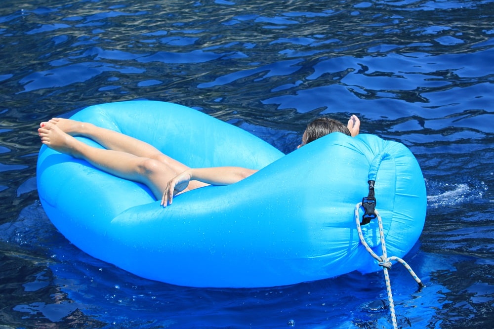 Eine Person entspannt sich auf einem blauen aufblasbaren Ring, der auf klarem, blauem Wasser in der Nähe einer Segelyacht schwimmt. Die Beine sind ausgestreckt, der Kopf ruht auf dem Rand des Rings.