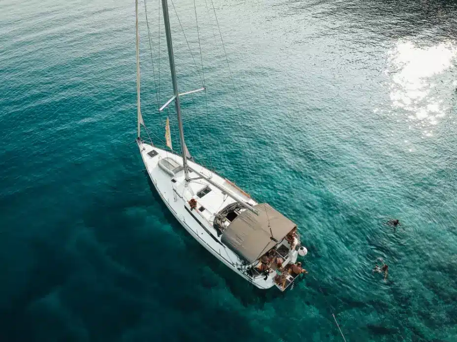 Luftaufnahme einer weißen Segelyacht auf klarem, türkisfarbenem Wasser mit Menschen, die sich an Deck entspannen und in der Nähe schwimmen. Das Sonnenlicht spiegelt sich im Wasser und verstärkt die ruhige Atmosphäre am Meer.