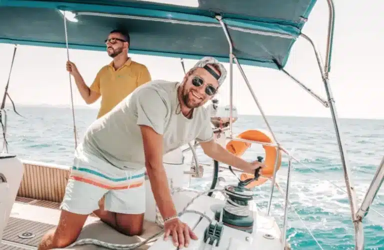 Zwei Männer auf einem Katamaran; einer mit Sonnenbrille lächelt in die Kamera und bedient Seile, der andere steuert im Hintergrund, um sie herum ein klares, blaues Meer und Himmel.