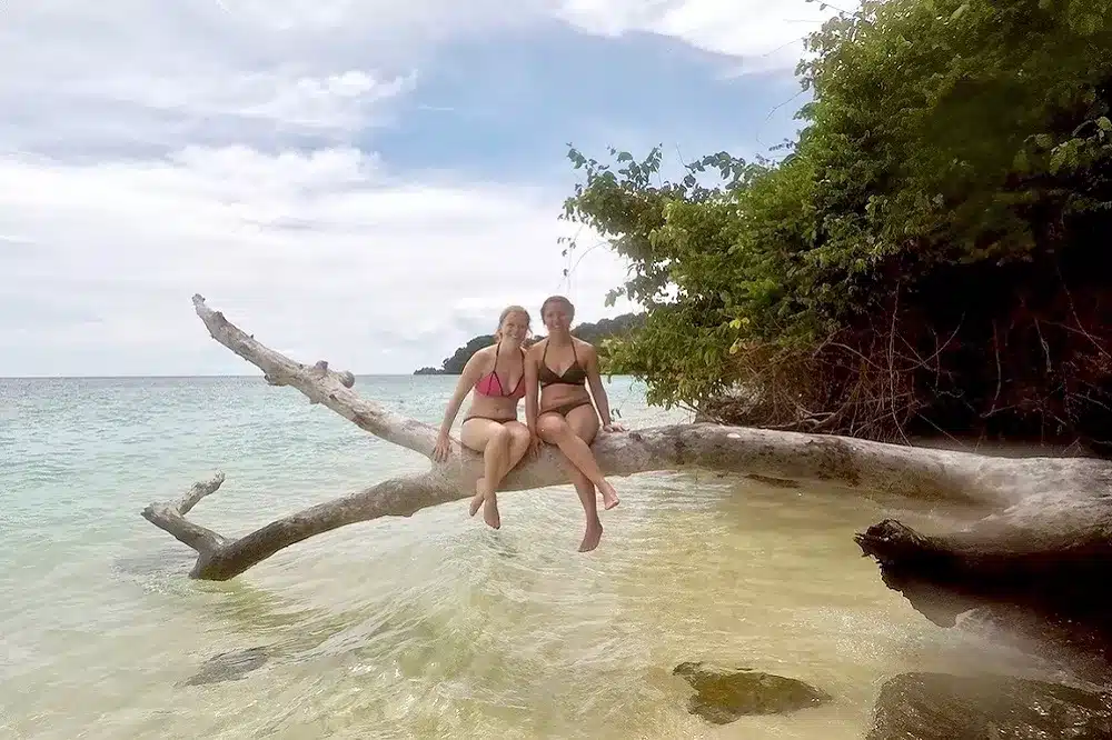 Zwei Menschen sitzen auf einem Ast über klarem Wasser an einem Strand, mit grünem Laub im Hintergrund und einem bewölkten Himmel darüber, und genießen den Blick auf die nahegelegenen Segelyachten.