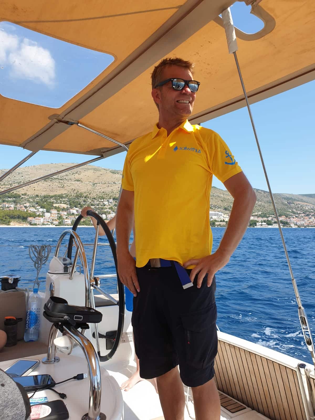 Ein Mann in einem gelben Poloshirt und schwarzen Shorts steht selbstbewusst auf einer Segelyacht, die Hände in die Hüften gestemmt, unter einem Baldachin, mit dem Meer und den Hügeln im Hintergrund.