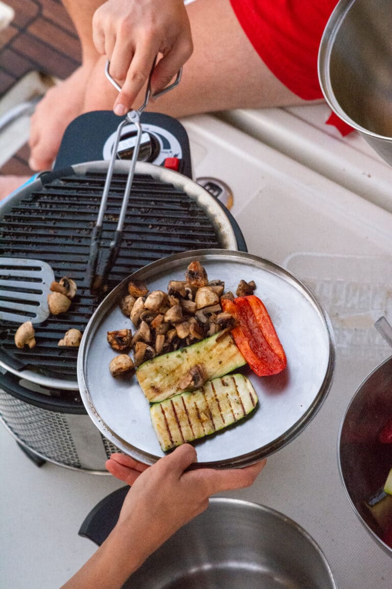 Eine Person grillt mit einer Zange Gemüse, darunter Pilze, Zucchinistreifen und eine rote Paprika, auf einem kleinen tragbaren Grill an Bord einer Segelyacht. Die Hände einer anderen Person halten eine Metallplatte, bereit, das gegrillte Gemüse aufzunehmen. Die Szene ist draußen auf Deck zu sehen, mit gemütlichen Sitzgelegenheiten während ihres Segelurlaubs.