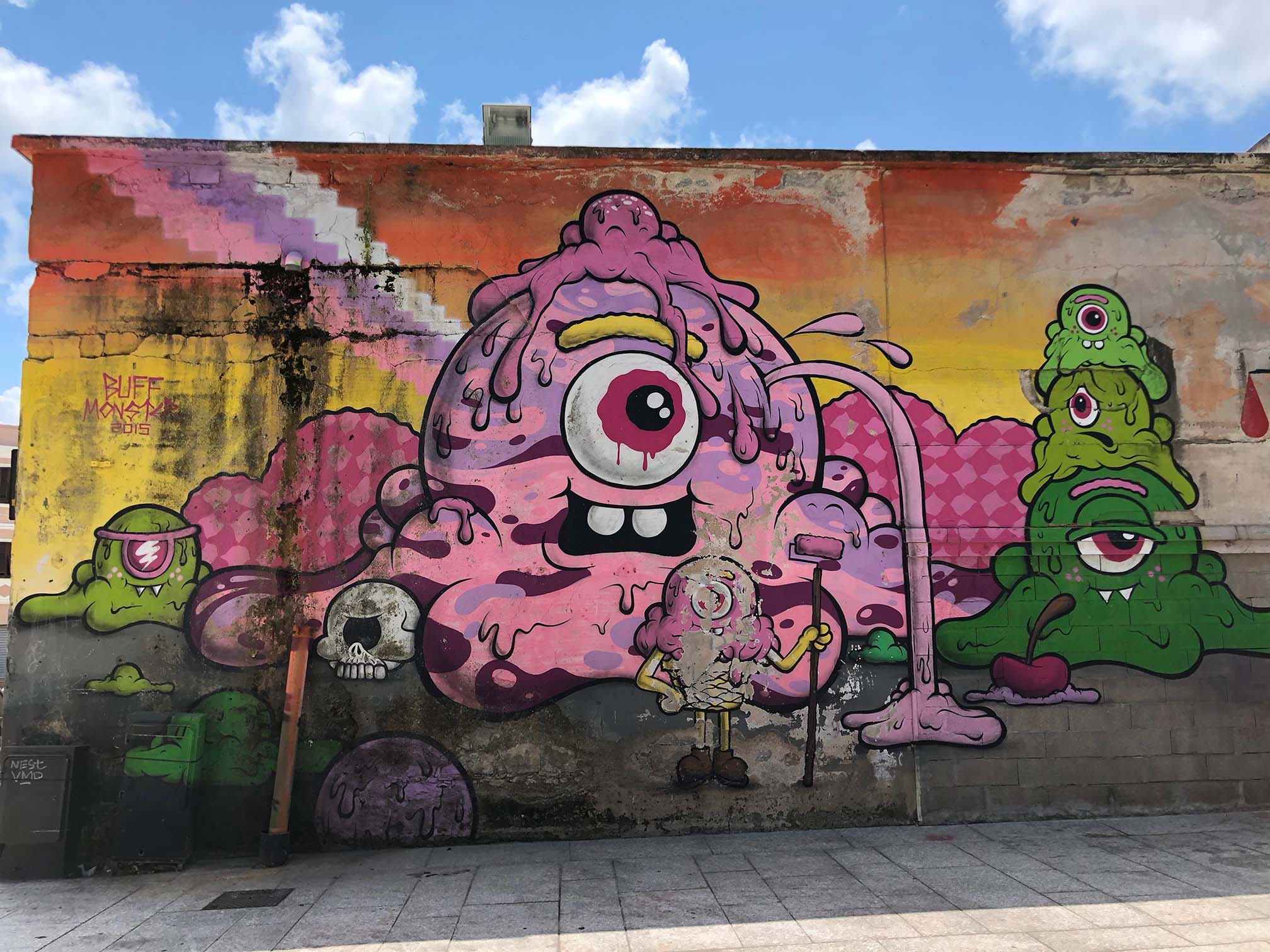 Ein farbenfrohes Street-Art-Wandbild zeigt in der Mitte eine große rosafarbene, einäugige Kreatur, von der Schleim tropft und die eine Farbrolle hält. Um sie herum sind weitere grüne und violette Monster, ein Totenkopf und abstrakte Formen zu sehen. Der helle Himmel darüber spiegelt einen lebhaften Katamaran-Segelurlaub mit vereinzelten Wolken wider.