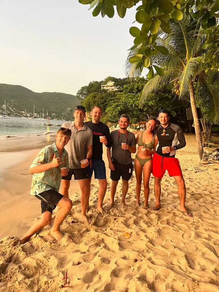 Eine Gruppe von sechs Personen, lächelnd und mit Getränken in der Hand, steht bei Sonnenuntergang an einem Sandstrand mit Palmen und Segelyachten im Hintergrund.