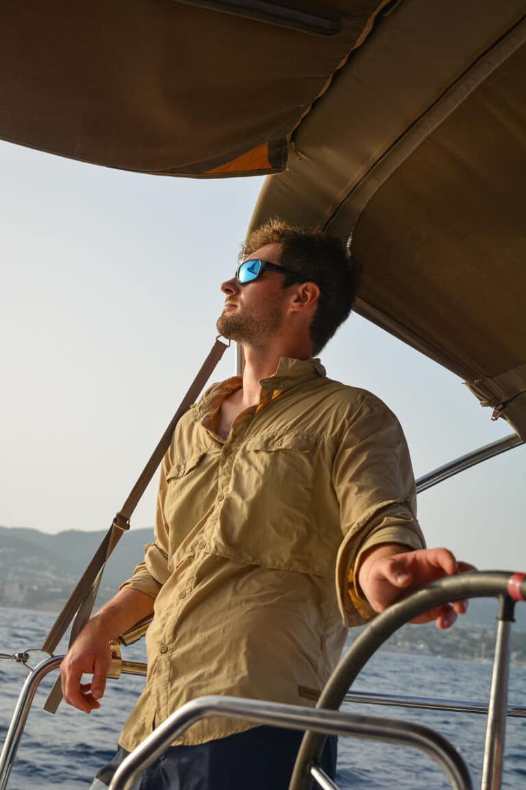 Ein Mann mit Sonnenbrille und beigem Hemd steuert mit konzentriertem Gesichtsausdruck ein Boot unter einem Baldachin auf einem Segeltörn mit dem Meer und den Hügeln im Hintergrund bei Sonnenuntergang.