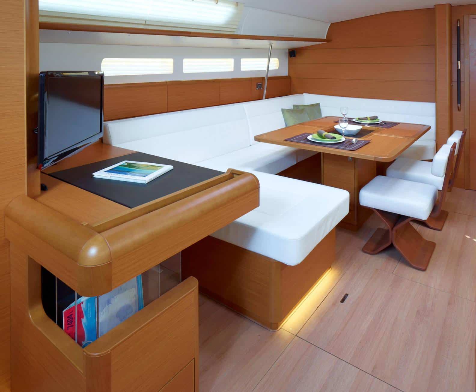 Innenansicht einer modernen Segelyachtkabine in elegantem Holzdesign mit weißem Sofa, passenden Stühlen und zwei Tischen mit Geschirr. Hell beleuchtet durch natürliches Licht aus den Seitenfenstern.
