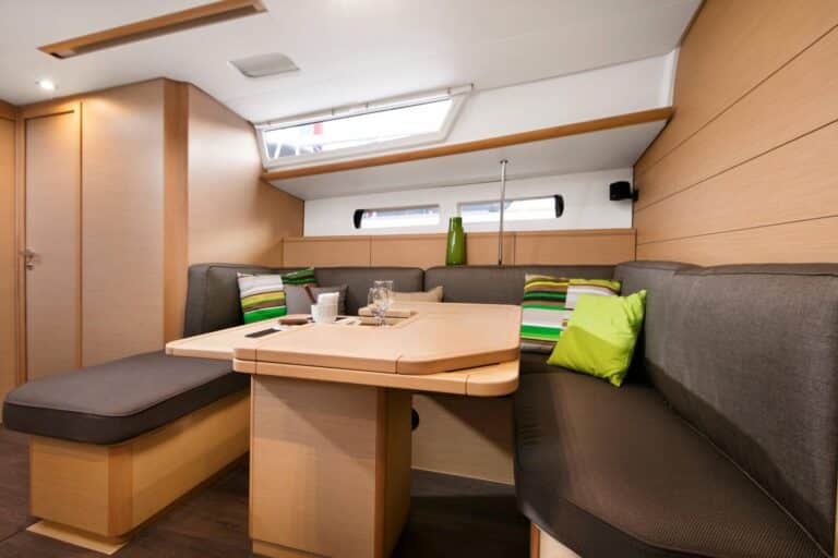 Innenansicht einer modernen Segelyachtkabine mit einem gemütlichen Essbereich mit einem Holztisch, umgeben von gepolsterten Bänken und hellgrünen Kissen. Natürliches Licht strömt durch ein Oberlicht herein.