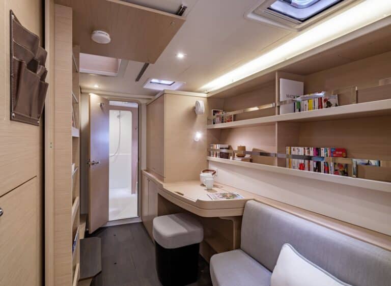 Innenansicht einer modernen Segelyachtkabine mit einer gemütlichen Sitzecke mit einem grauen Sofa, einem Holzschreibtisch, eingebauten Bücherregalen voller Bücher und stilvoller Deckenbeleuchtung. Eine Tür führt in einen anderen Raum.