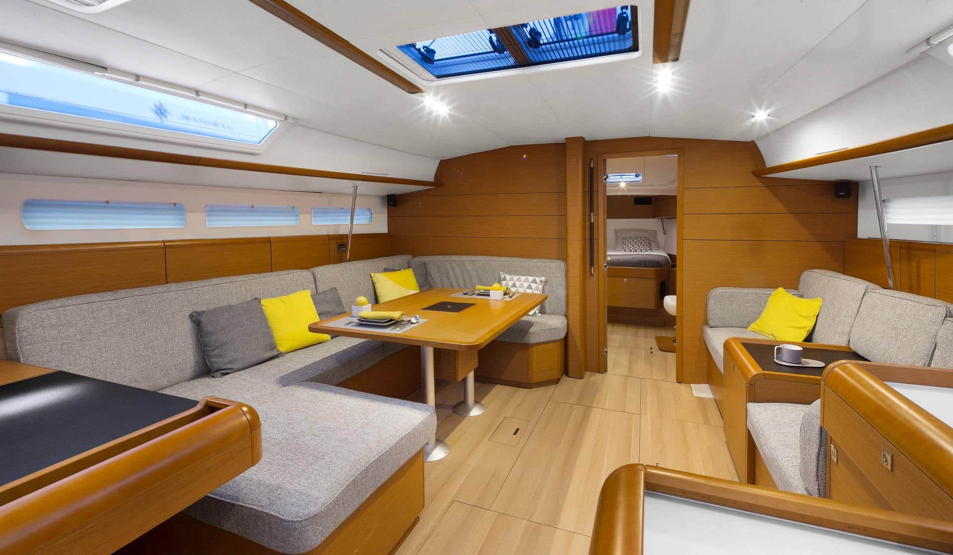 Innenraum einer modernen Segelyacht mit stilvollem Sitzbereich mit grauen Sofas, einem Holztisch mit Büchern und gelben Akzentkissen. Helles Licht durch Oberlichter und Seitenfenster, mit warmer Holzvertäfelung.